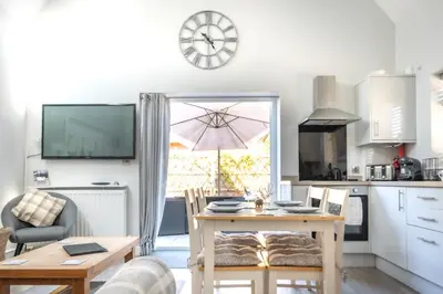 a living room with a table and a kitchen with a clock at Hedgehog Cottage in Bury Saint Edmunds