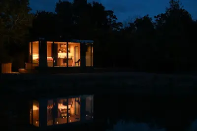 a glass house at night with a reflection in the water at Mirror Houses Pound Farm - Pond OOD in Saxmundham