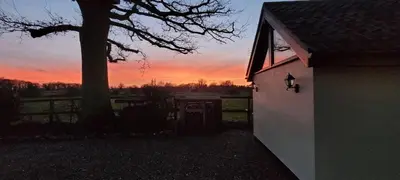 a sunset behind a house with a tree and a fence at The Acorn - Roudham Norwich in Norwich