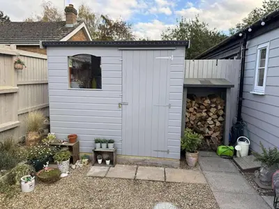 a grey shed with a pile of fire wood at Cosy Flint Cottage in Thurston