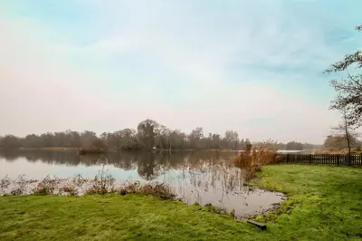 a view of a lake with grass and a fence at Heronsmead, sleeps 8 - Large Thorpeness house on the banks of Thorpness Meare - a boating lake - Close to Thorpeness beach, Golf Club and Thorpeness Country Club - Aldeburgh Coastal Cottages in Thorpeness