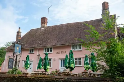 a pink building with tables and green umbrellas at Cosy and eco-friendly cabins next to 16th century pub in Newmarket